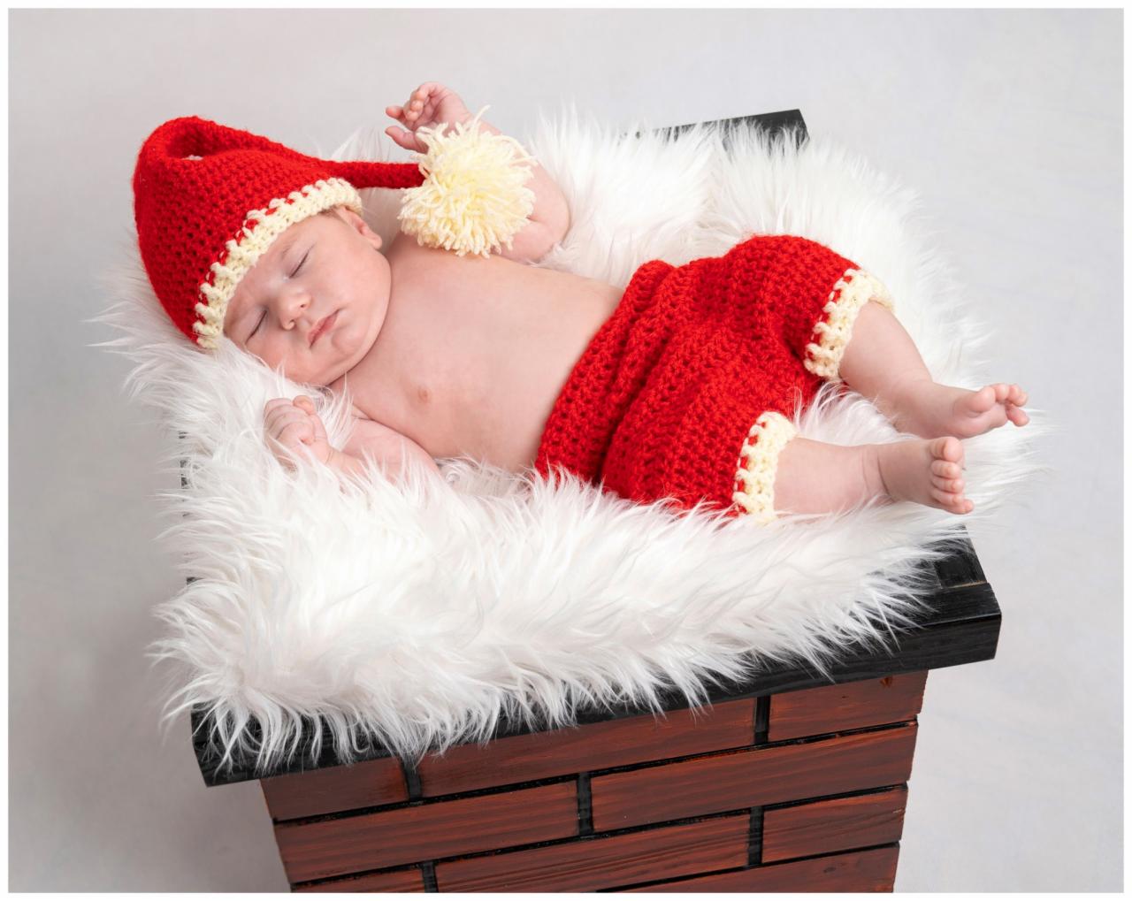Śpiące niemowlę w czerwonym, dzierganym stroju Mikołaja, leżące na białym, puszystym futerku w drewnianej skrzyni imitującej komin.