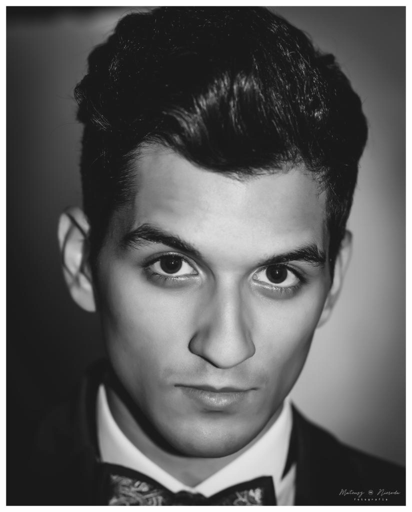 Czarno-biały portret młodego mężczyzny w muszce, z wyraźnym spojrzeniem i klasyczną fryzurą, oświetlony z boku.