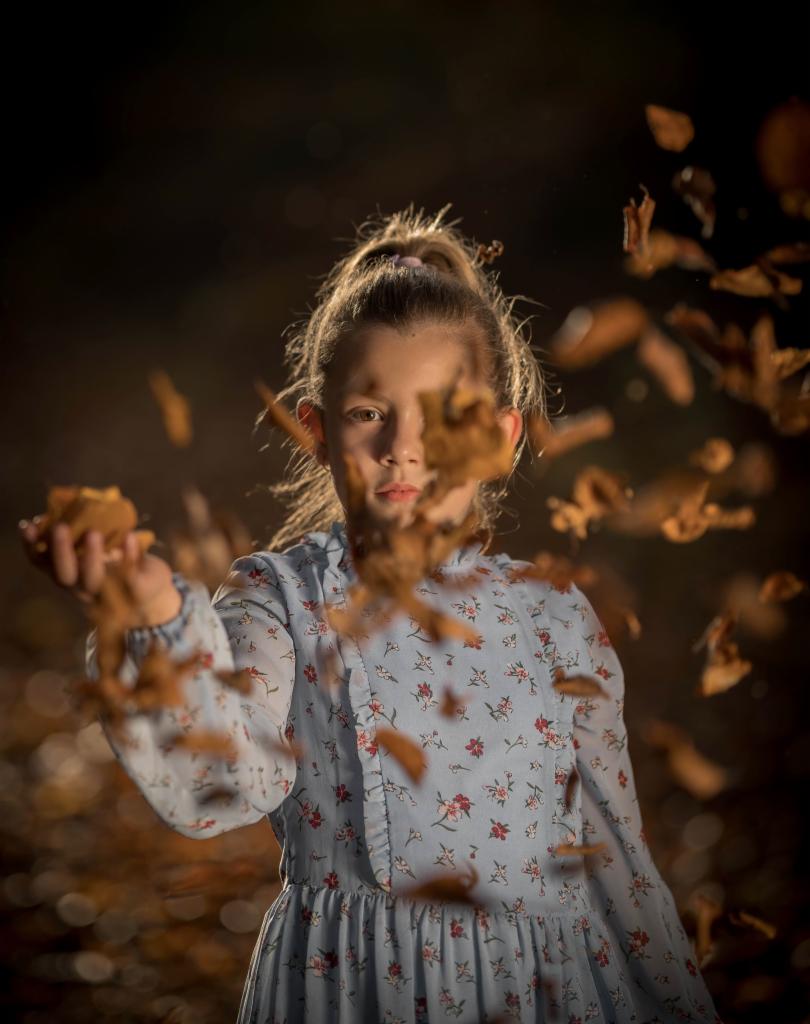 Dziewczynka w kwiecistej sukience rzuca w górę jesienne liście, oświetlona ciepłym światłem na ciemnym tle.