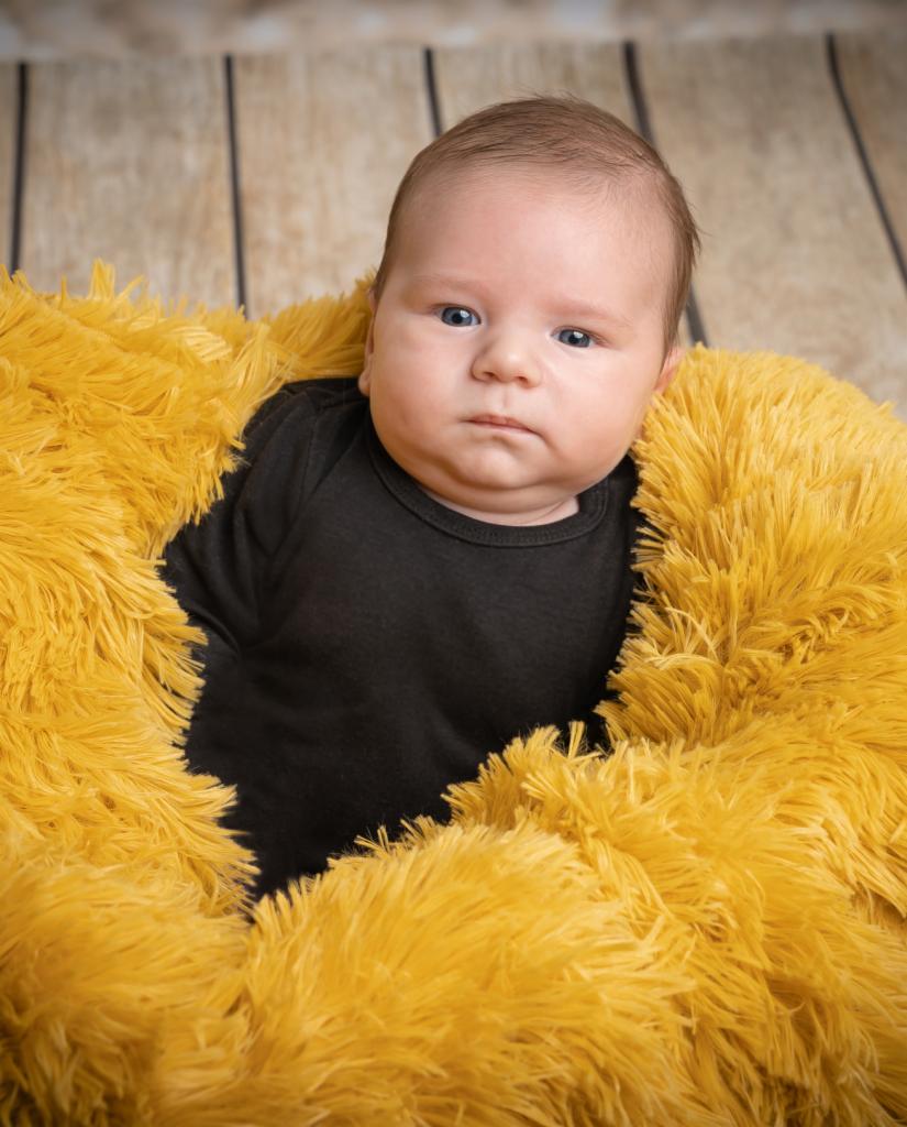 Niemowlę w czarnym body leży na puszystym, żółtym kocyku na tle drewnianej podłogi.