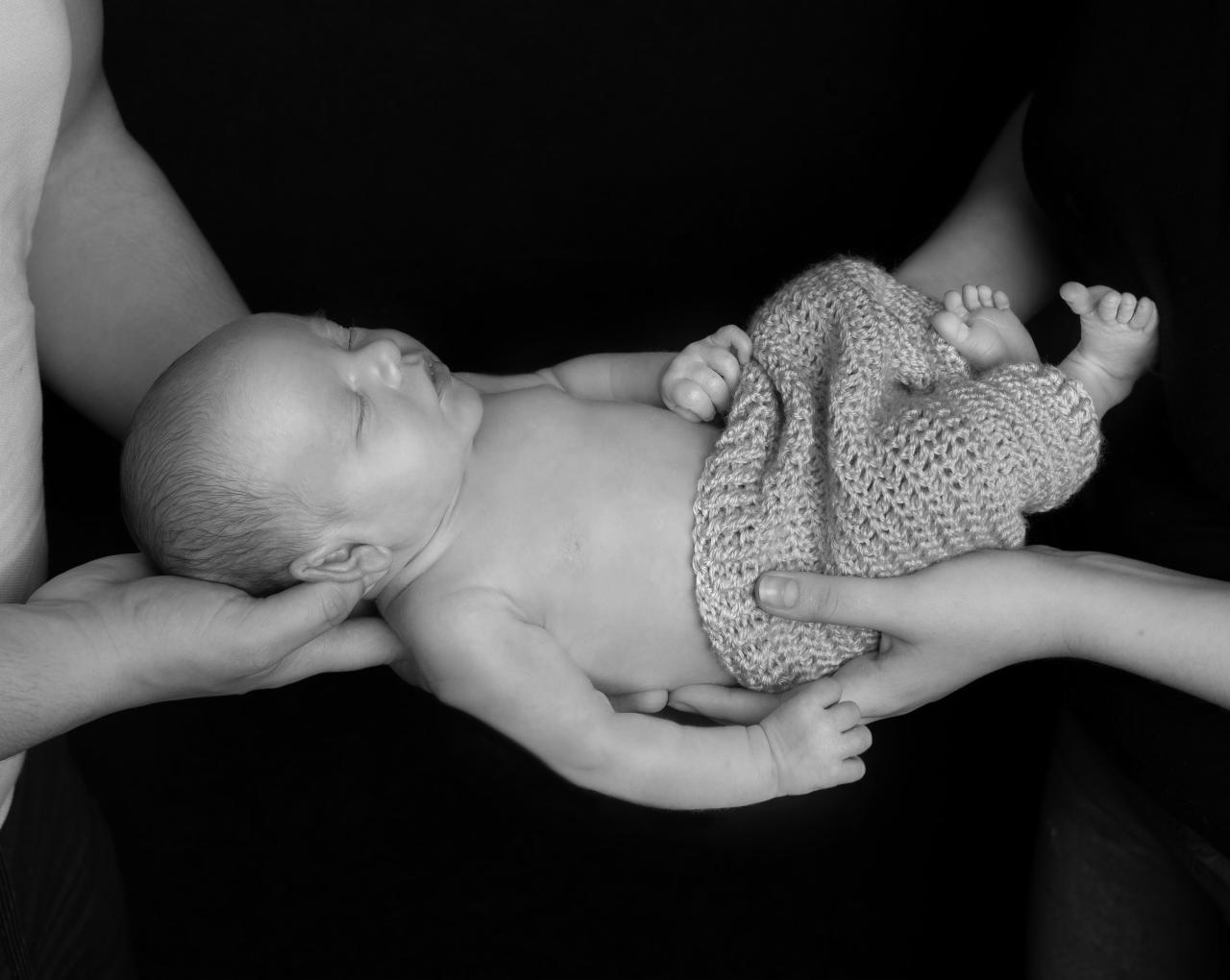 Czarno-białe zdjęcie noworodka śpiącego w dłoniach rodziców, owiniętego w dziergany kocyk.