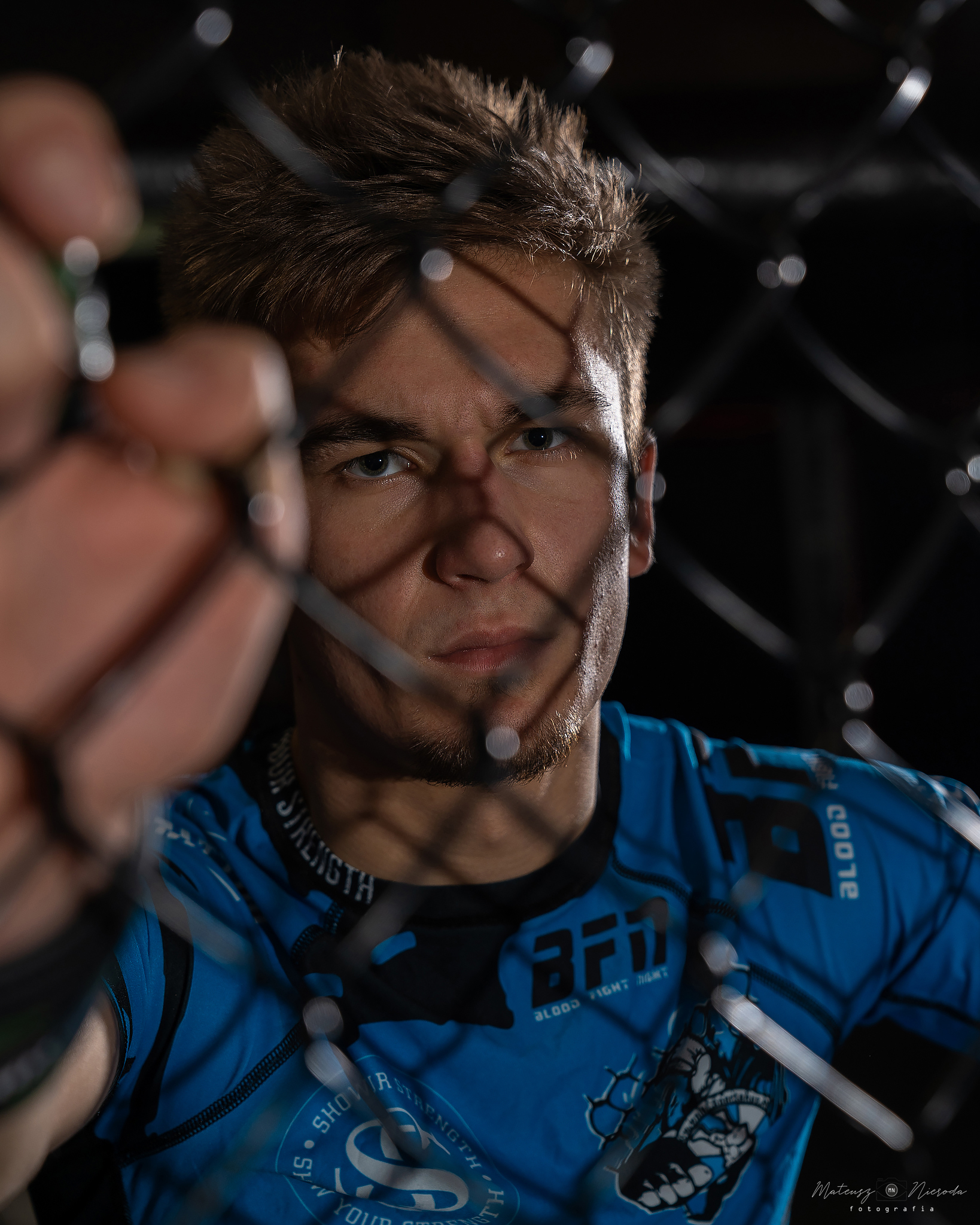 Portret młodego mężczyzny w niebieskiej koszulce sportowej z nadrukami, stojącego za klatką z siatki, z poważnym wyrazem twarzy i skupionym wzrokiem.