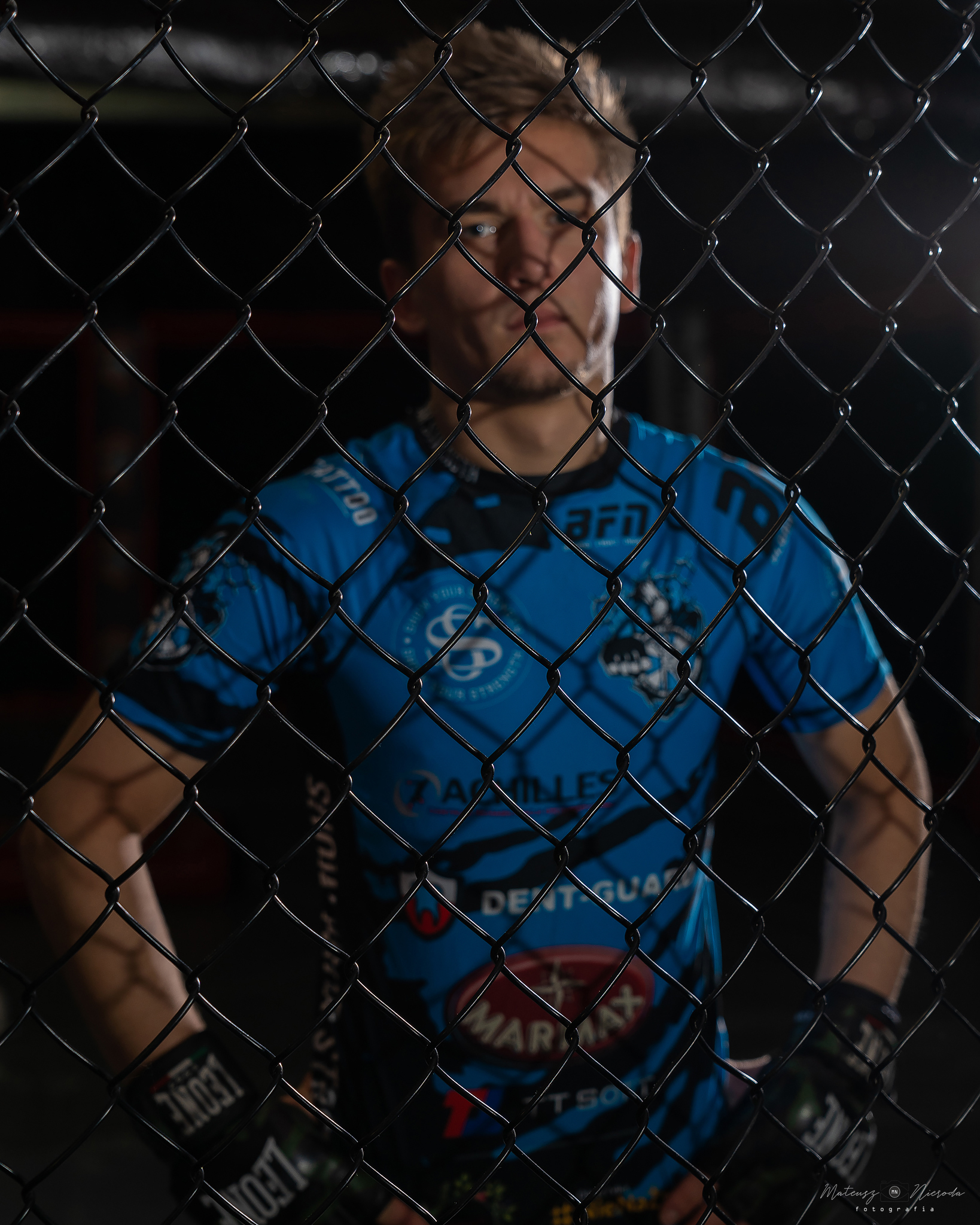 Zawodnik MMA w niebieskiej koszulce z logo sponsorów, widziany przez siatkę klatki.