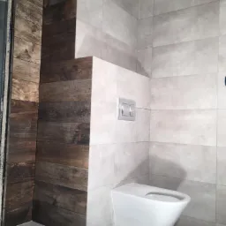 Wykończona łazienka z drewnopodobnymi i jasnoszarymi płytkami, wisząca toaleta z chromowanym przyciskiem spłukiwania.