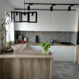 Nowoczesna kuchnia z białymi szafkami, drewnianym blatem i szarymi płytkami imitującymi marmur, oświetlona czarnymi lampami sufitowymi i wiszącymi lampami z żarówkami Edison.