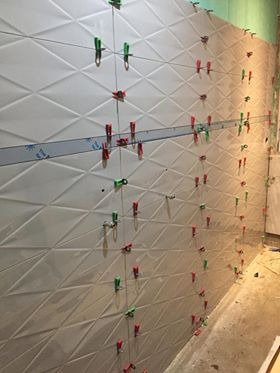 Ściana z białych, trójwymiarowych płytek ceramicznych ułożonych w romby, z widocznymi systemami poziomowania płytek (klipsy i kliny) oraz sznurkami wyznaczającymi linie, w trakcie prac...