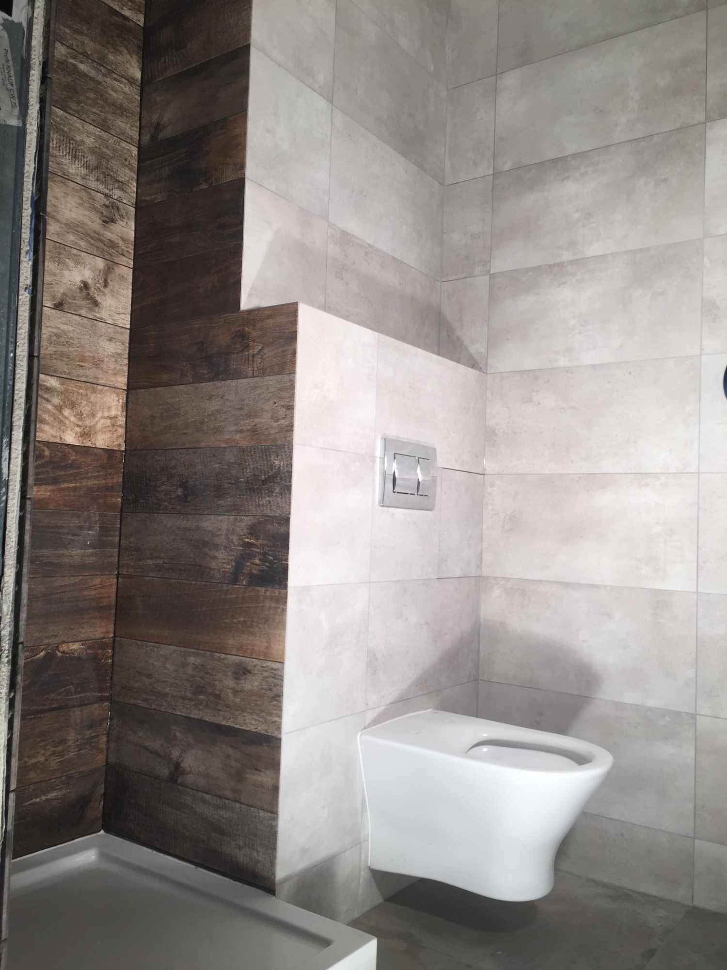 Wykończona łazienka z drewnopodobnymi i jasnoszarymi płytkami, wisząca toaleta z chromowanym przyciskiem spłukiwania.