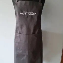 Szary fartuch kuchenny z białym napisem 'Piekarnia i Cukiernia POD STRZECHĄ' zawieszony na czarnym manekinie na tle białej ściany.