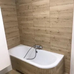 Biała wanna obudowana płytkami imitującymi drewno, z chromowaną baterią, na ścianach i podłodze płytki w podobnym stylu, widok pod kątem.