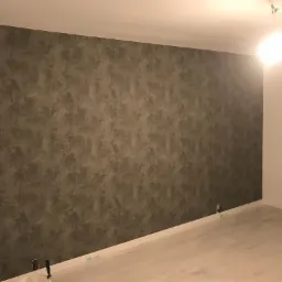 Ściana z dekoracyjną strukturą w odcieniach szarości, po zakończeniu prac, widoczne gniazdka elektryczne przy podłodze i fragment okna.