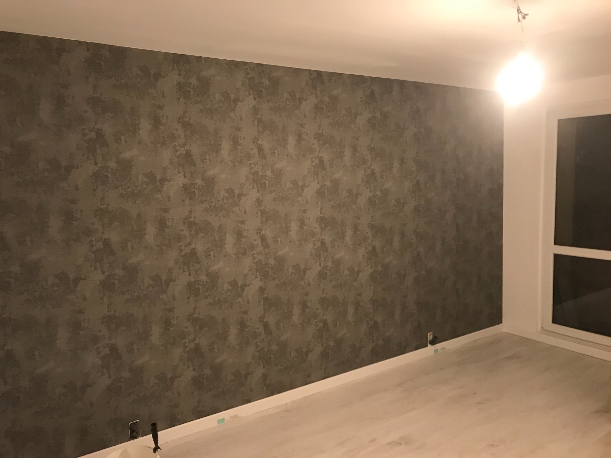 Ściana z dekoracyjną strukturą w odcieniach szarości, po zakończeniu prac, widoczne gniazdka elektryczne przy podłodze i fragment okna.