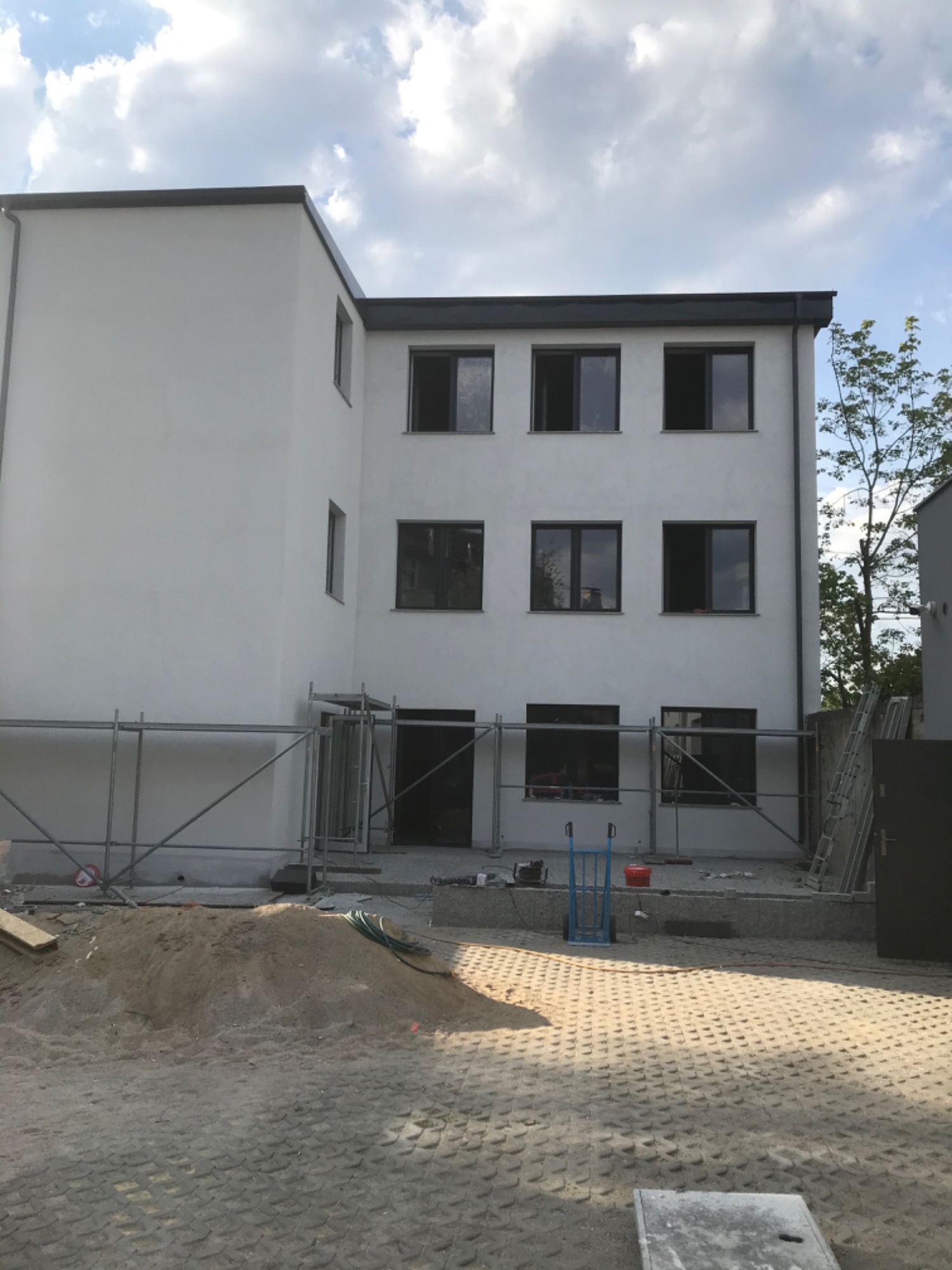 Nowo wybudowany, biały budynek mieszkalny z ciemnymi ramami okiennymi, częściowo otoczony rusztowaniami, z widocznym stosem piasku i materiałów budowlanych na pierwszym planie.