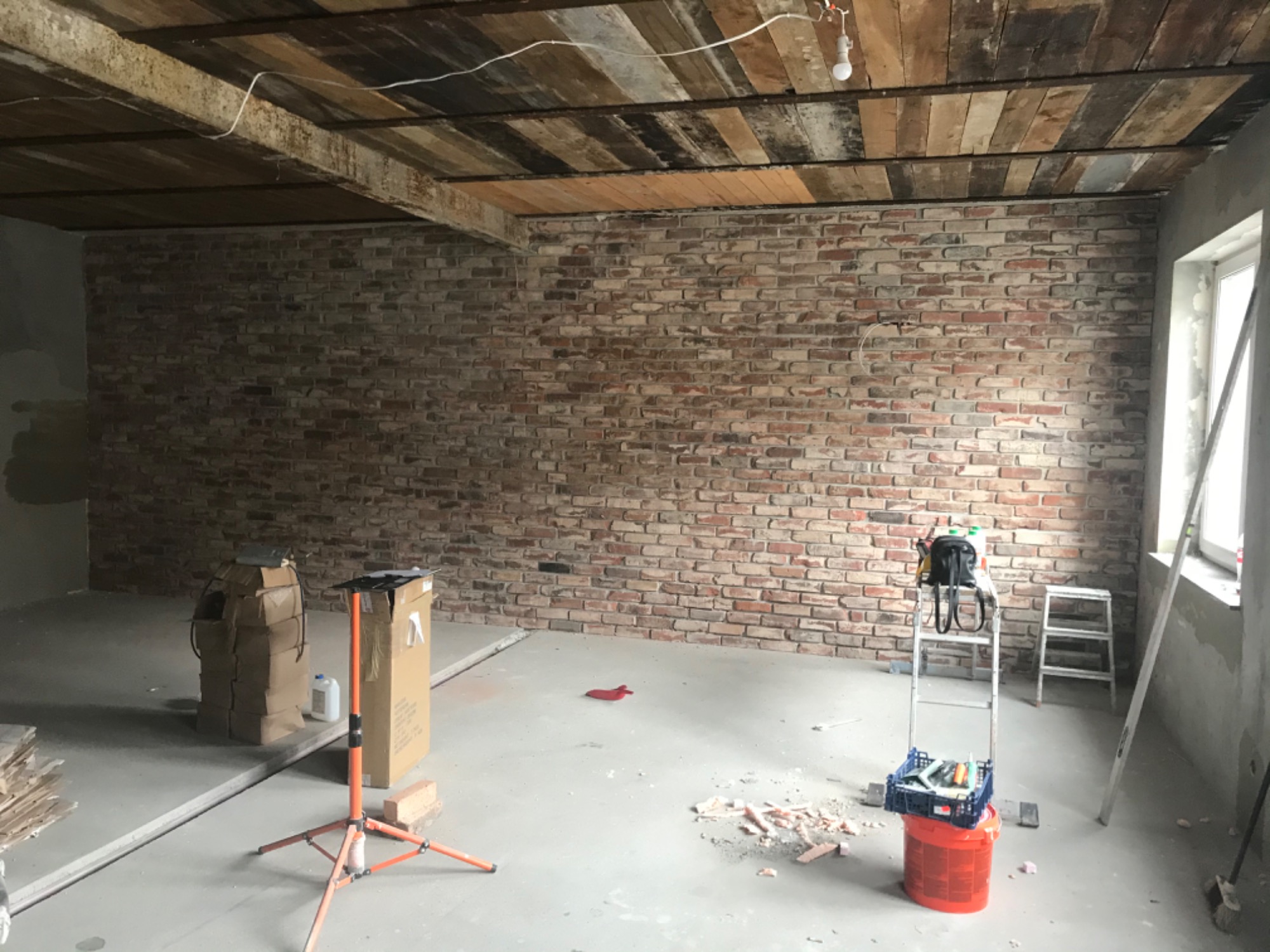 Surowe wnętrze w trakcie prac: ceglana ściana, drewniany sufit z widocznymi belkami, drabina z narzędziami, kartony i resztki materiałów budowlanych na podłodze.