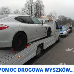 Białe Porsche Panamera na lawecie pomocy drogowej w Wyszkowie, zabezpieczone pomarańczowymi pasami, w tle radiowóz policyjny i kobieta stojąca na chodniku z wymalowanymi białymi gołębiami.