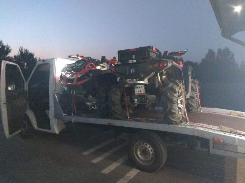 Biała laweta z otwartymi drzwiami kabiny przewozi dwa quady z dużymi kołami, zabezpieczone pasami transportowymi. Jeden z quadów ma czarny kufer na bagażniku.
