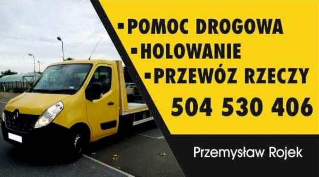 Żółta laweta Renault z czarnymi elementami, na tle szaro-żółtej reklamy z napisem 'Pomoc Drogowa, Holowanie, Przewóz rzeczy' i numerem telefonu.