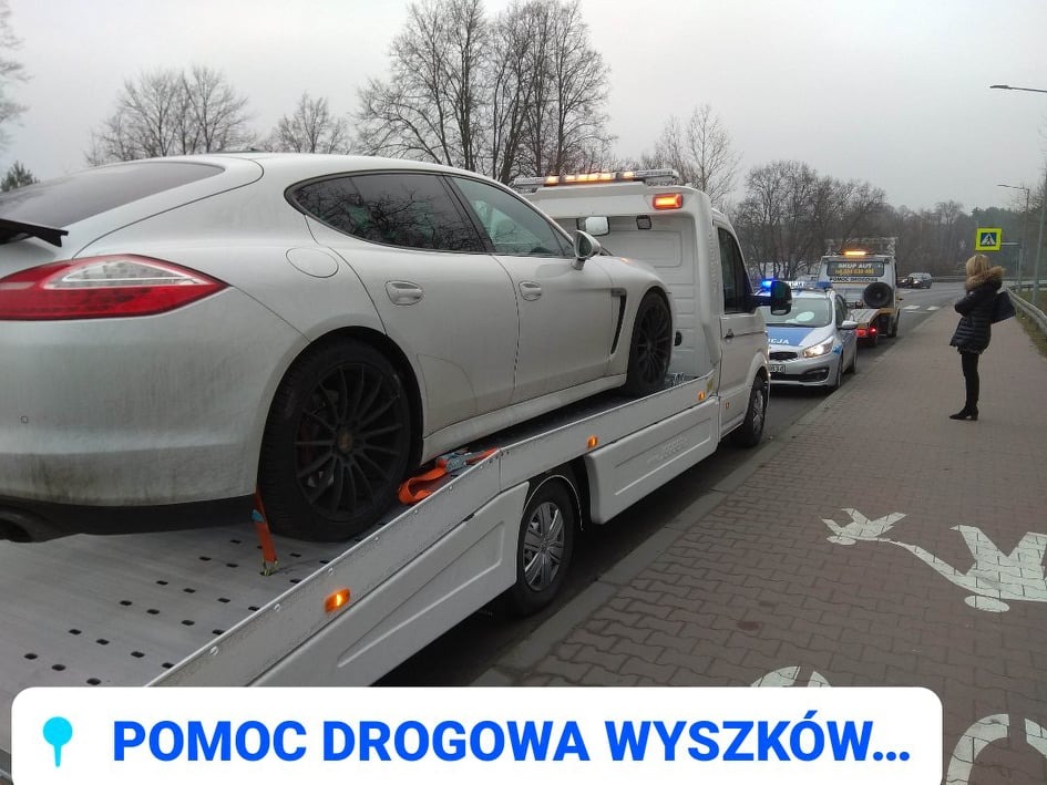 Białe Porsche Panamera na lawecie pomocy drogowej w Wyszkowie, zabezpieczone pomarańczowymi pasami, w tle radiowóz policyjny i kobieta stojąca na chodniku z wymalowanymi białymi gołębiami.