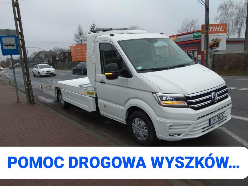 Biała laweta Volkswagen Crafter na tle ulicy miejskiej z widocznym znakiem przystanku autobusowego i budynkiem z reklamą STIHL, zaparkowana na chodniku.