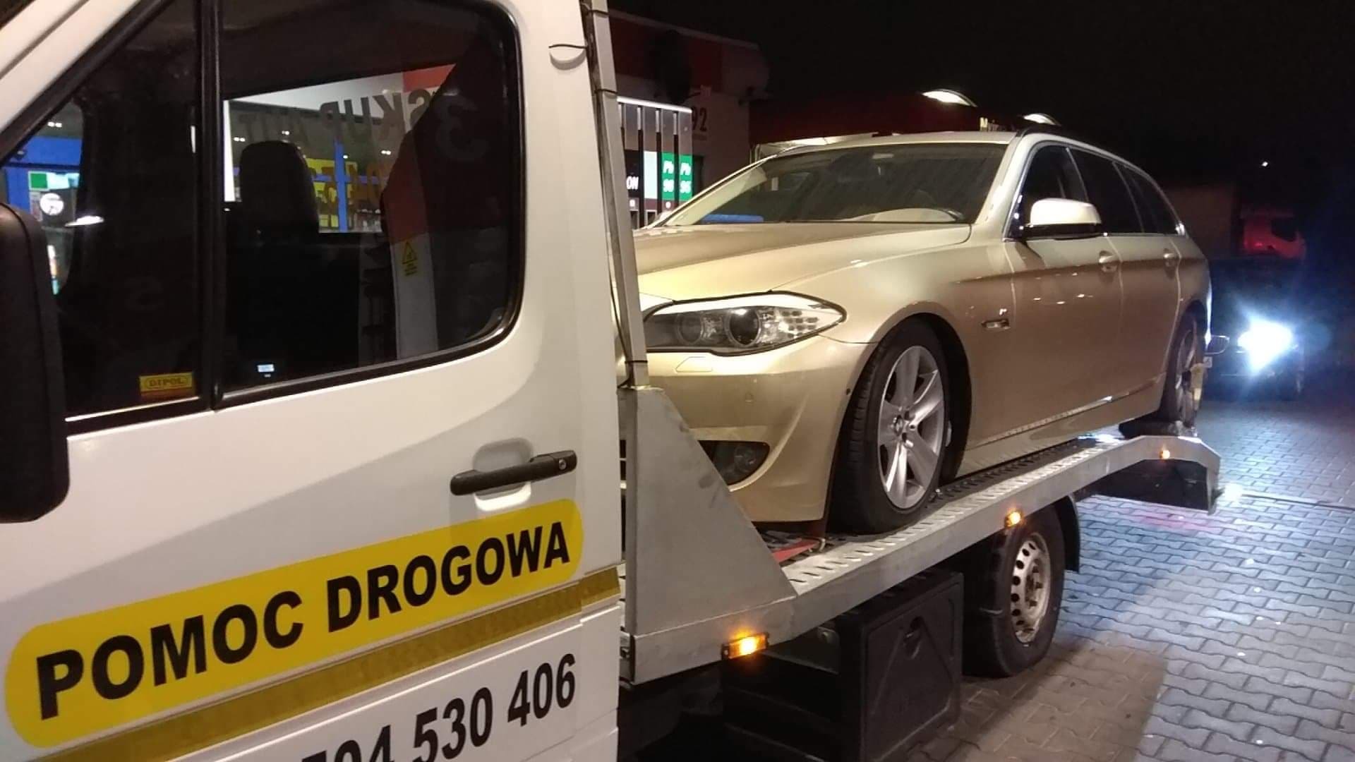 Złoty samochód osobowy BMW kombi na lawecie pomocy drogowej z żółtym napisem POMOC DROGOWA, na tle stacji benzynowej nocą.
