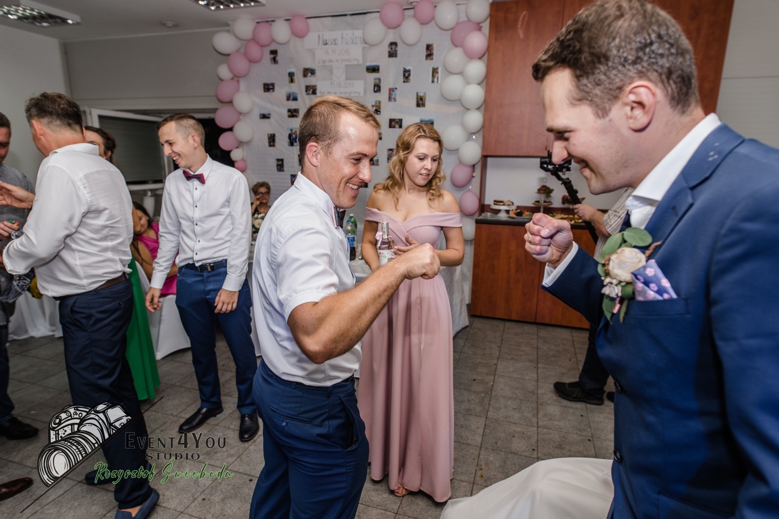 Uśmiechnięci panowie w eleganckich strojach wymieniający się 'żółwikami' na tle dekoracji balonowych i tablicy ze zdjęciami podczas wesela.