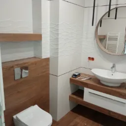 Nowoczesna łazienka z drewnianymi akcentami: umywalka nablatowa, wisząca toaleta, białe płytki z falistym wzorem, drewniane półki i okrągłe lustro z podświetleniem.