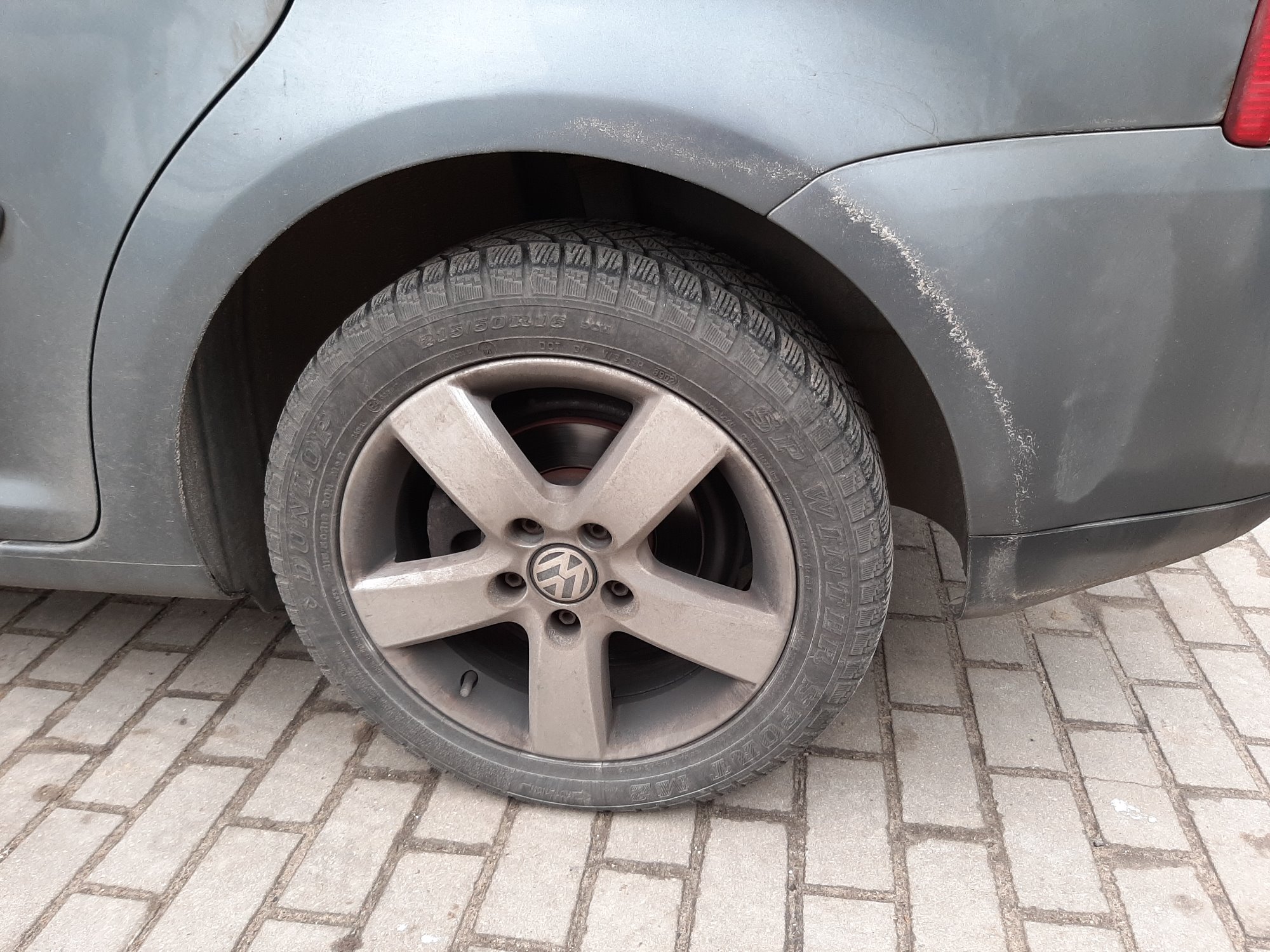 Szare auto z widocznymi zabrudzeniami na karoserii i feldze, zaparkowane na kostce brukowej.