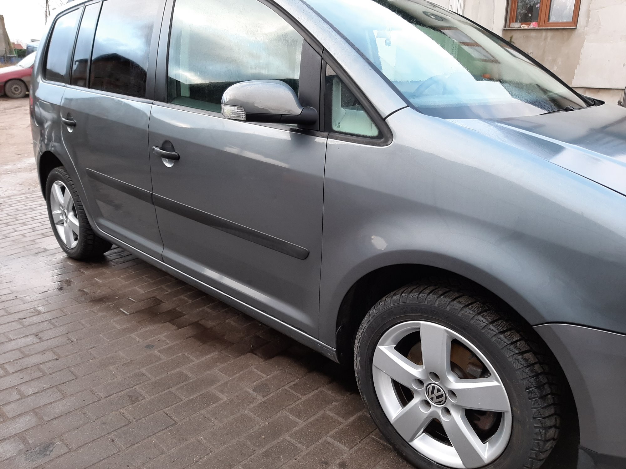 Szary samochód osobowy marki Volkswagen, widziany z boku, zaparkowany na mokrej, brązowej kostce brukowej. Widoczne detale: felgi aluminiowe, lusterko boczne z kierunkowskazem, fragment karoserii...