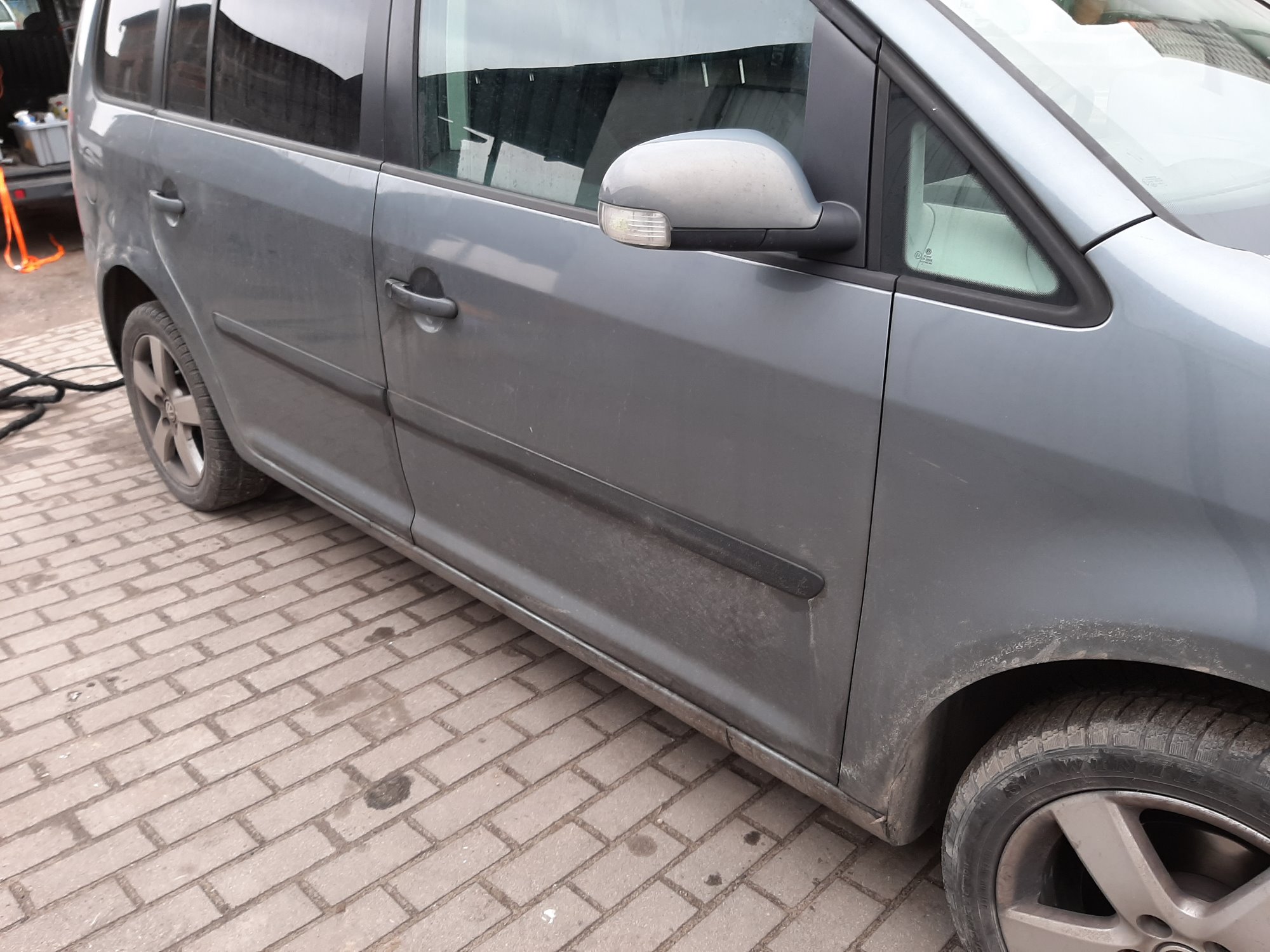 Szary, brudny samochód typu minivan zaparkowany na chodniku z kostki brukowej, widoczny z boku, z otwartym bagażnikiem w tle.