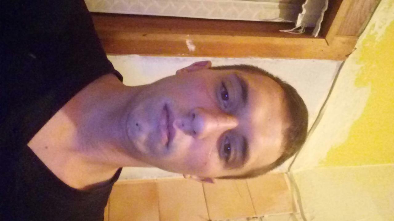 Mężczyzna z bliska na tle ściany w trakcie malowania, widoczne niedomalowane fragmenty i drewniana rama okienna. Nietypowy kadr z góry.