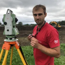 Pomiar geodezyjny w terenie: mężczyzna z niwelatorem Leica na statywie, w tle wykop i koparka. Czerwona koszulka z logo Leica. Pochmurne niebo.