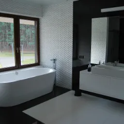 Nowoczesna łazienka z wolnostojącą wanną przy oknie z widokiem na las, biało-czarne płytki podłogowe i umywalka na szafce.