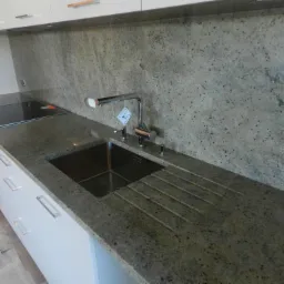 Kuchnia z blatem kamiennym w odcieniach szarości i beżu, z wbudowanym zlewem i ociekaczem, bateria z etykietą, płyta indukcyjna w tle, pod szafkami wiszącymi.