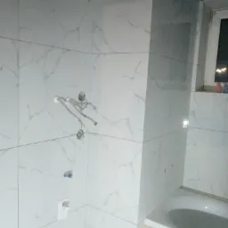 Ściany łazienki wyłożone białymi płytkami imitującymi marmur, widoczne rury instalacji wodnej przed montażem armatury, plastikowe krzyżyki dystansowe w fugach oraz fragment wanny.