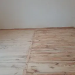 Podłoga z desek sosnowych, część po renowacji z widocznym usłojeniem, druga część przed renowacją, surowa, jasna, w rogu pomieszczenia.