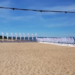 Plaża z rzędem białych flag reklamowych z logo sponsor&oacute;w wydarzenia, widok na morze i błękitne niebo, dekoracyjne lampki na sznurze wiszące nad piaskiem.