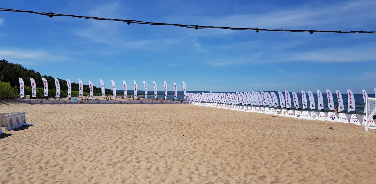 Plaża z rzędem białych flag reklamowych z logo sponsorów wydarzenia, widok na morze i błękitne niebo, dekoracyjne lampki na sznurze wiszące nad piaskiem.