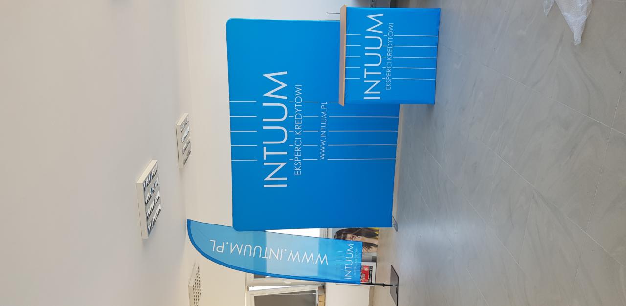 Niebieskie stoisko promocyjne z logo firmy 'INTUUM' i adresem strony internetowej, ustawione na szarych płytkach podłogowych, z widoczną flagą reklamową i fragmentem okna w tle.