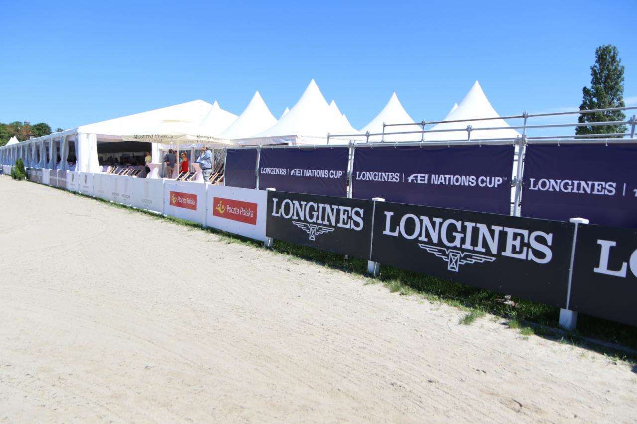 Widok na teren zawodów jeździeckich z białymi namiotami w tle, ogrodzony panelami reklamowymi z logo Longines i Poczty Polskiej.