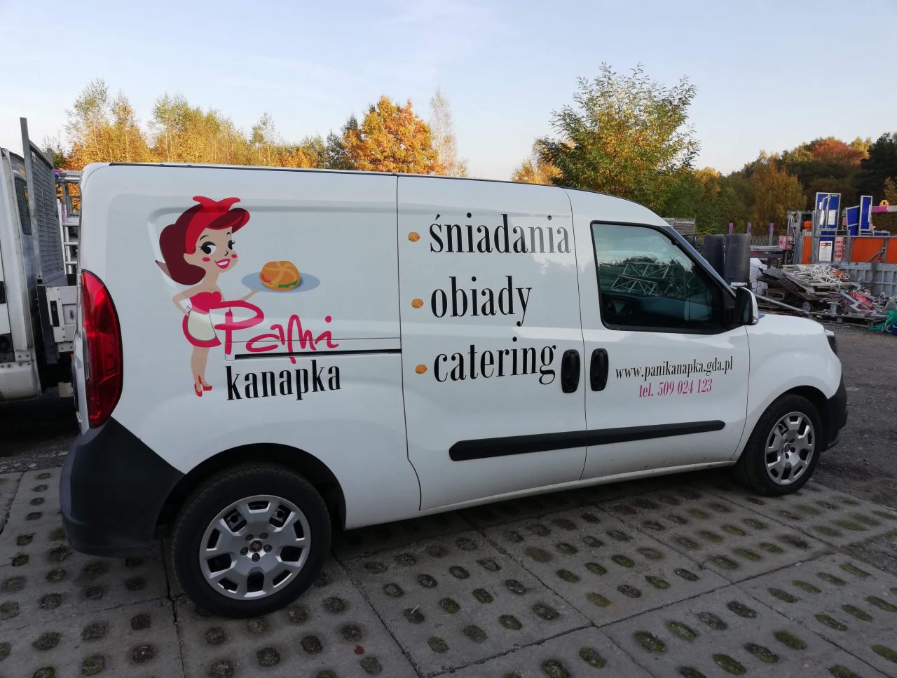 Biały samochód dostawczy z grafiką przedstawiającą postać kobiety z kanapką, napisy 'śniadania', 'obiady', 'catering' oraz adres strony internetowej i numer telefonu firmy oferującej usługi...