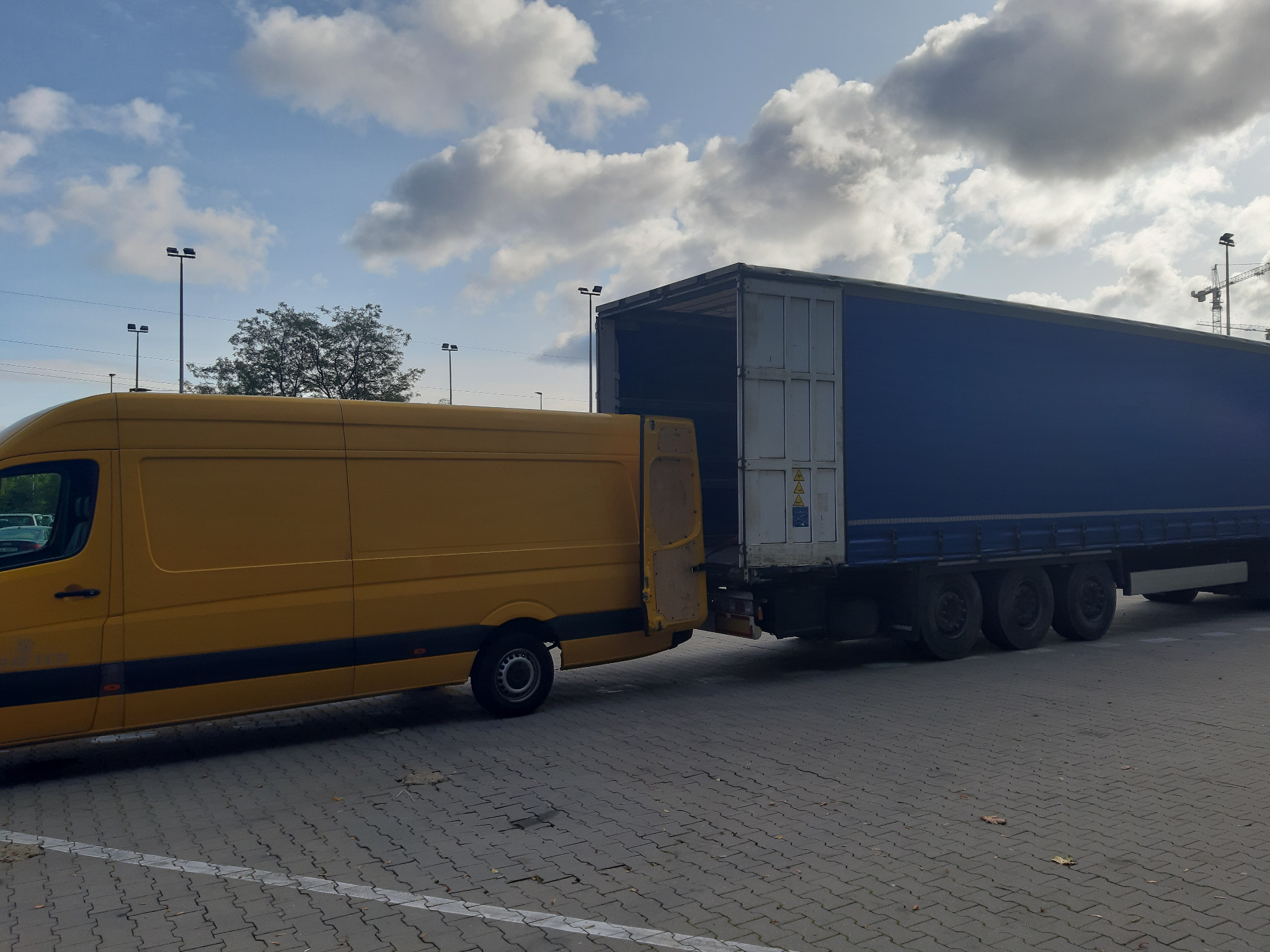 Żółty bus dostawczy z otwartymi tylnymi drzwiami zaparkowany przy naczepie ciężarówki z otwartymi drzwiami w przestrzeni miejskiej z widocznymi latarniami i żurawiami w tle.