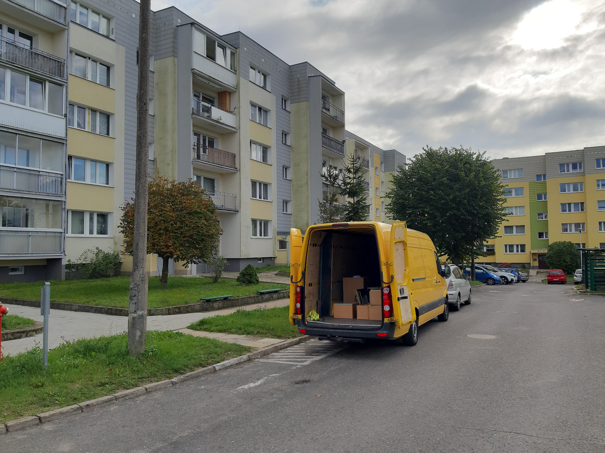 Żółty bus z otwartymi tylnymi drzwiami, zaparkowany na osiedlowej uliczce, z widocznymi kartonami wewnątrz, na tle bloków mieszkalnych i zaparkowanych samochodów.