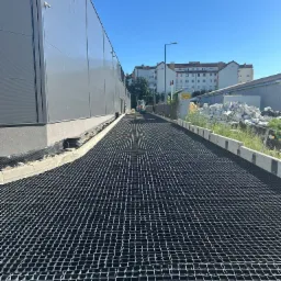 Utwardzona droga z kratą, widok z perspektywy, budynek przemysłowy w tle, błękitne niebo. Utwardzenie terenu z zastosowaniem geokraty.