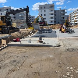 ZiemneUslugi.pl Jakub Szczudło - Prace brukarskie na terenie budowy osiedla: fragment ułożonej kostki, ekipa z maszyną, koparka Volvo, palety z kostką, gruz i piasek. W tle budynki mieszkalne.