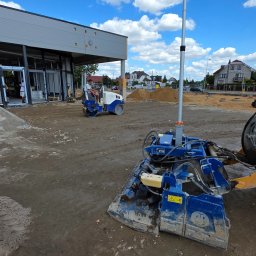 ZiemneUslugi.pl Jakub Szczudło - Utwardzanie terenu przed budynkiem komercyjnym. Widoczny walec drogowy i ubijarka wibracyjna. W tle hałda piasku i zabudowa miejska w słoneczny dzień.