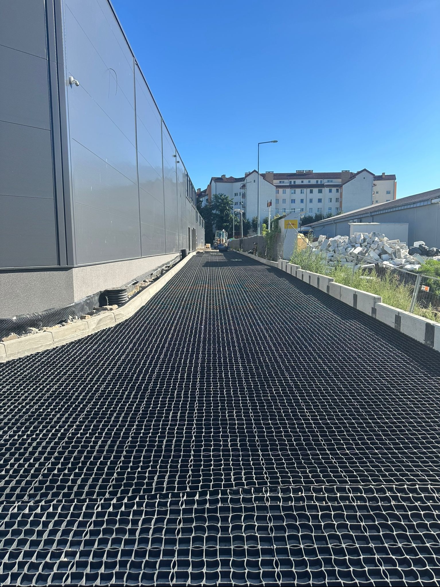 Utwardzona droga z kratą, widok z perspektywy, budynek przemysłowy w tle, błękitne niebo. Utwardzenie terenu z zastosowaniem geokraty.