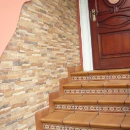 Schody wejściowe wyłożone brązowymi płytkami z dekoracyjnym wzorem, ściana z imitacji kamienia i drewniane drzwi.