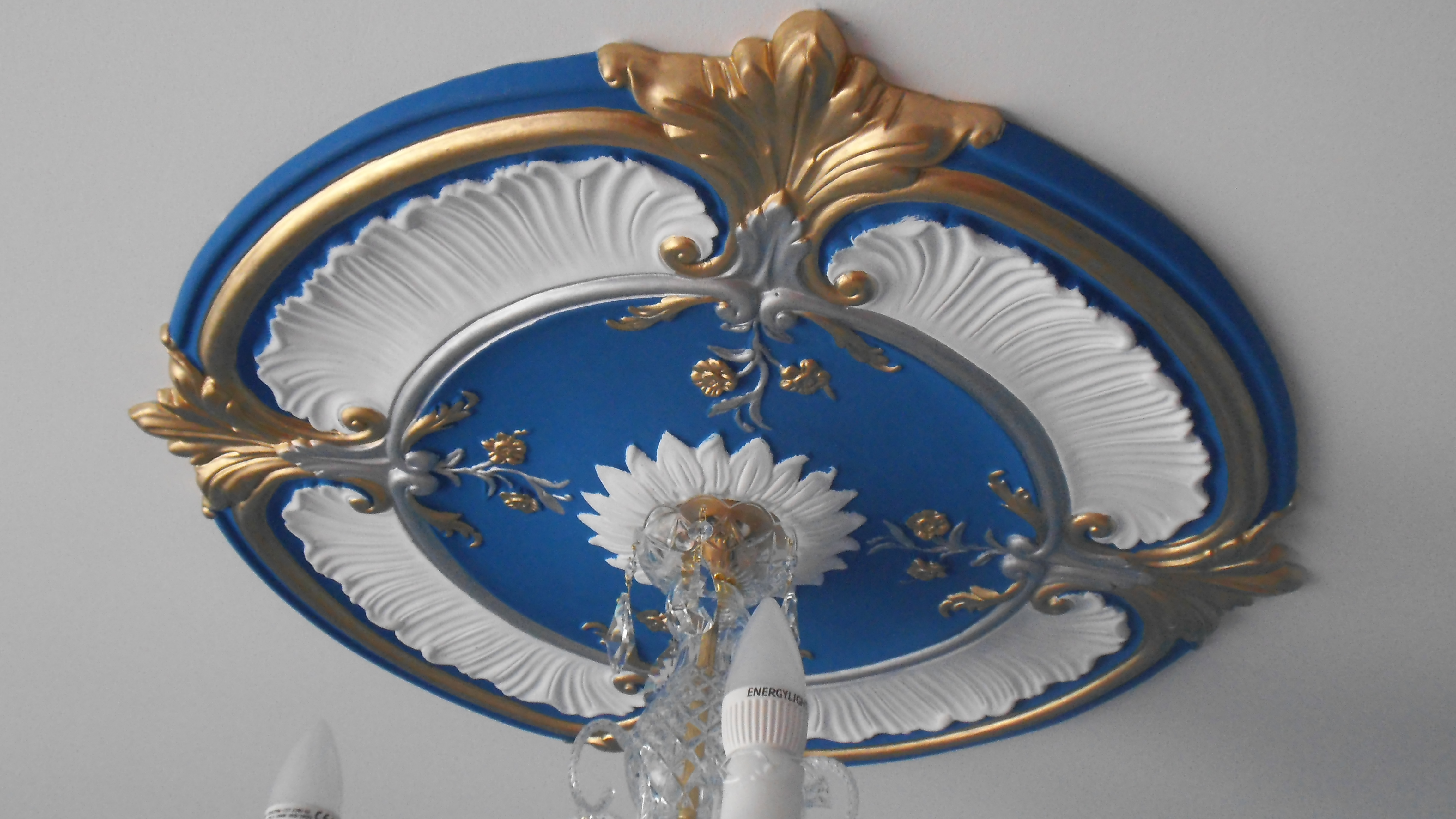 Ozdobny, okrągły rozetowy element dekoracyjny sufitu w kolorze niebieskim, złotym i białym, z kryształowym żyrandolem w centrum, oświetlonym żarówkami LED.