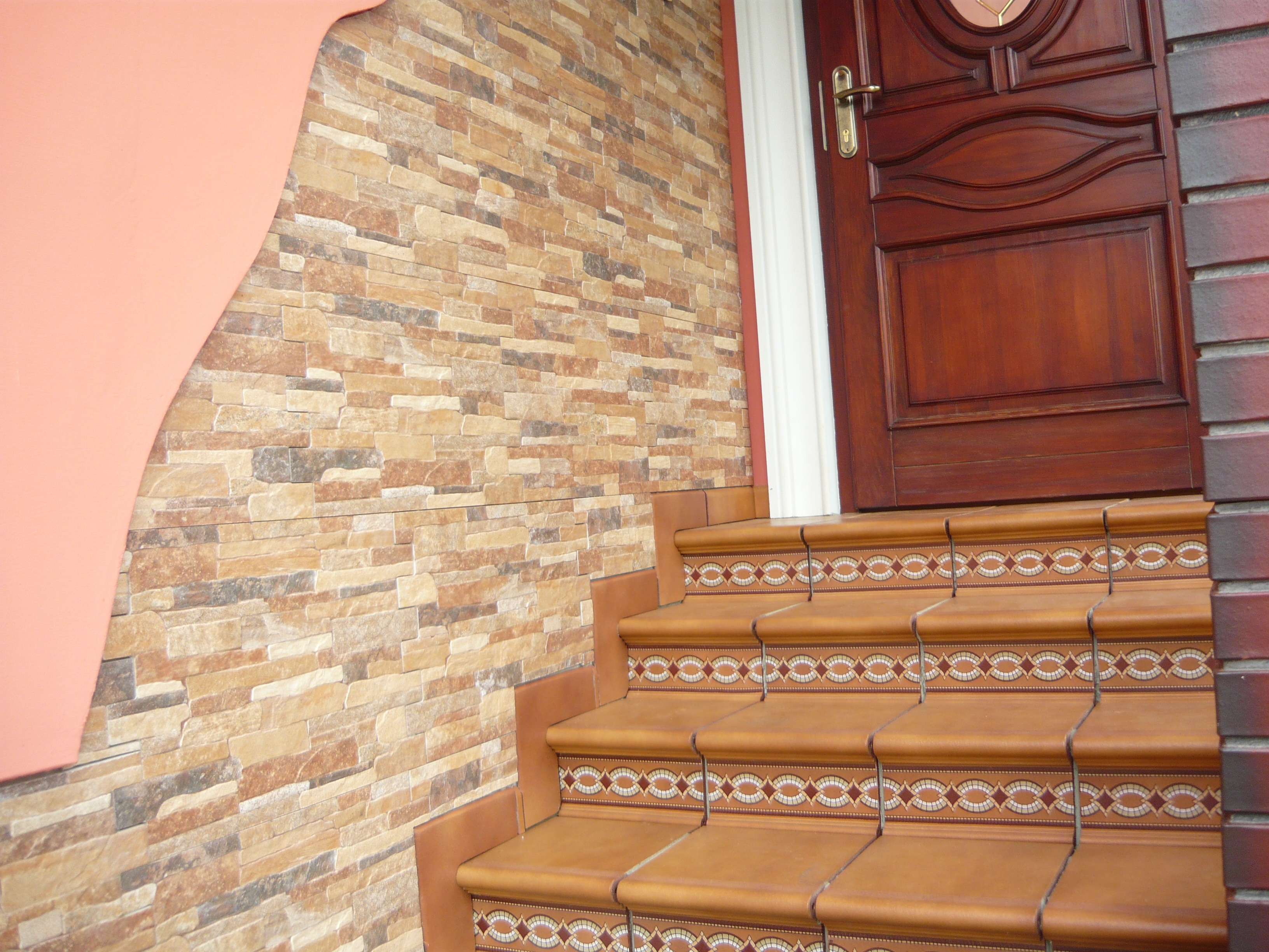 Schody wejściowe wyłożone brązowymi płytkami z dekoracyjnym wzorem, ściana z imitacji kamienia i drewniane drzwi.