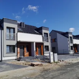 Nowoczesne szeregowe domy w trakcie wykańczania elewacji, z widocznymi fragmentami ocieplenia i przygotowanym podłożem pod chodnik z kostki brukowej, błękitne niebo w tle.