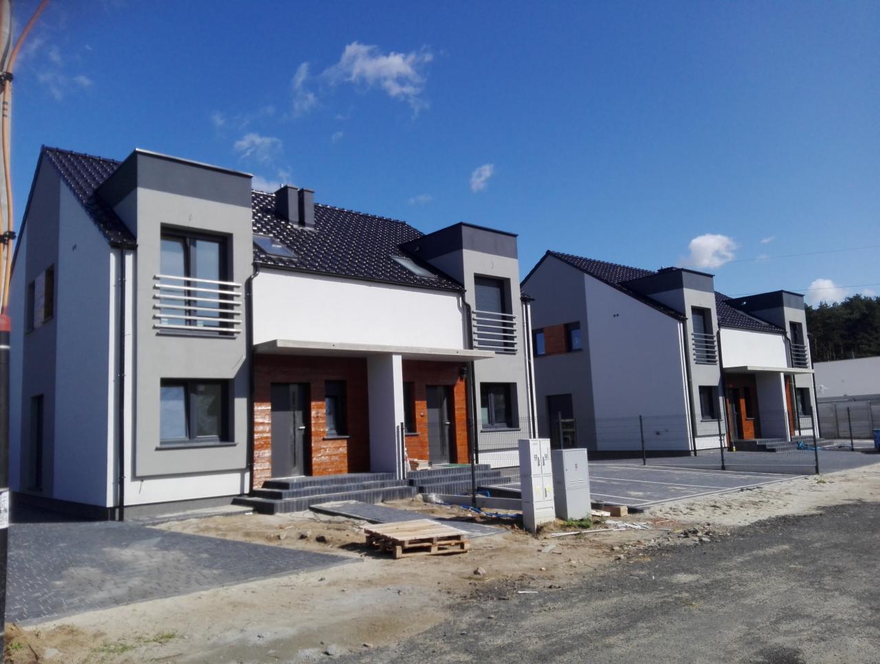 Nowoczesne szeregowe domy w trakcie wykańczania elewacji, z widocznymi fragmentami ocieplenia i przygotowanym podłożem pod chodnik z kostki brukowej, błękitne niebo w tle.